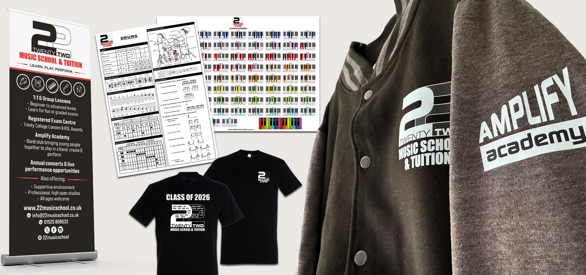 Promotional collage for Amplify Academy and 22 music school and tuition, showing branded apparel and printed materials: a dark gray varsity jacket with white logo, a black T-shirt, a black sweatshirt, a drum sheet and a piano sheet, and a roller banner on a light grey background.
