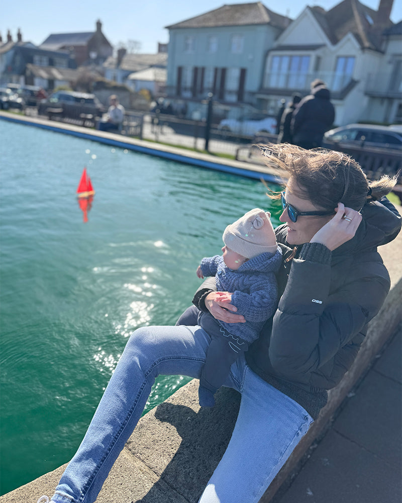 FFS Print and Media founder, Emma Denmark, sitting on a stone ledge by a lake holds a baby. The woman wears a dark puffer jacket, light blue jeans, and white sneakers, while the baby wears a light coloured beanie and a blue knit outfit.